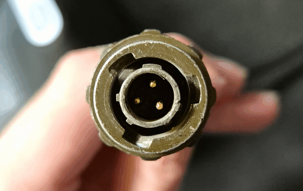 A top down view of a worn-down connector with three pins in the center. The casing is in a dark military green.
