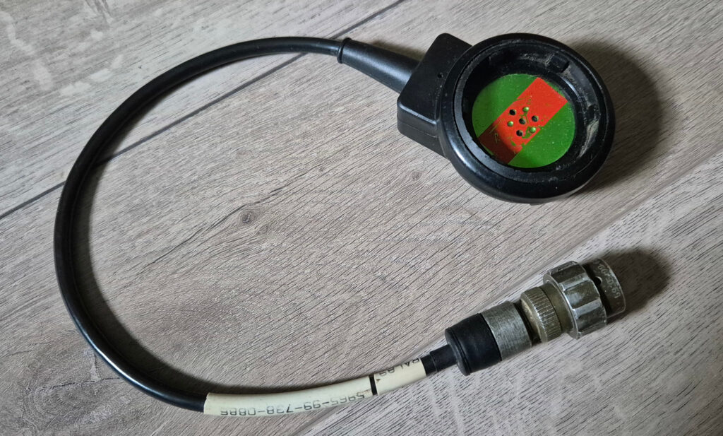 A clansman microphone module with a short wire attached to it. The microphone module is made from black plastics, the membrane is green with a red stripe across.